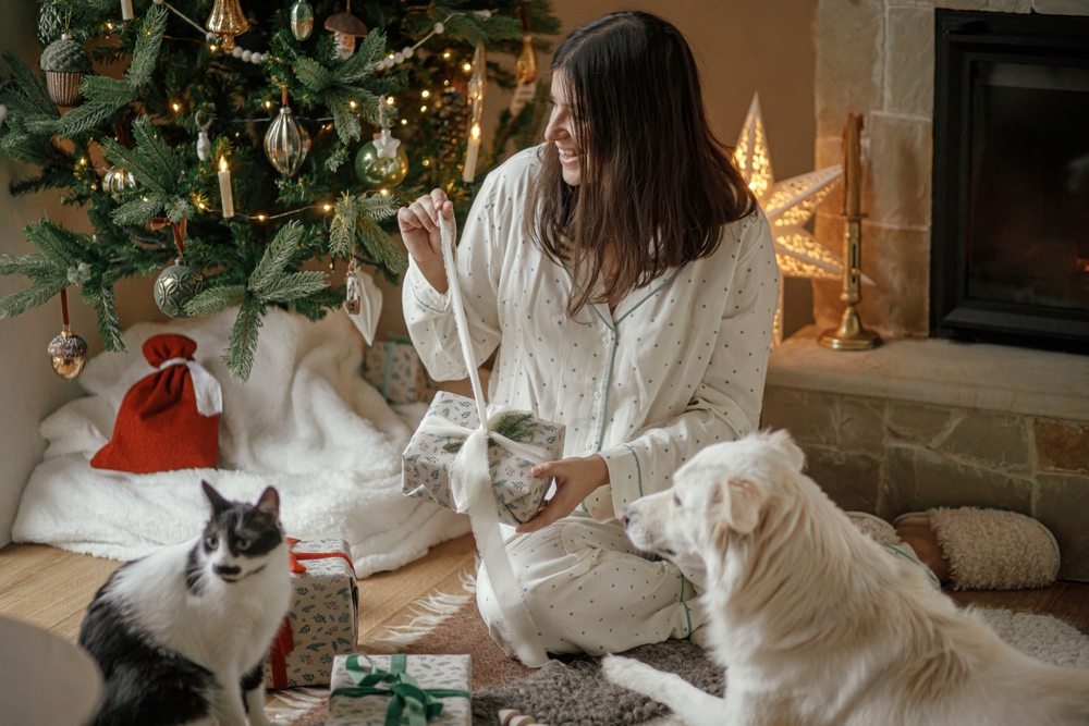 A woman in pajamas sits beside a Christmas tree, smiling as she interacts with a cat and a dog. Presents and a glowing star ornament are visible.