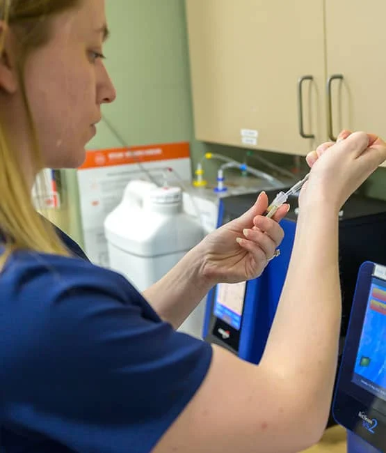 A female veterinarian in a blue uniform preps a syringe at a workstation with medical equipment in the background.