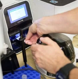 Close-up of hands using veterinary laboratory equipment, including a small digital monitor and a glass container, to conduct scientific experiments.