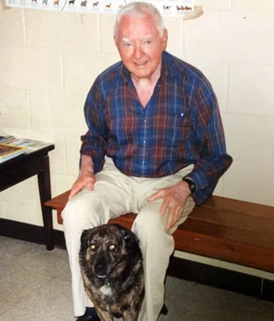 Elderly man with a warm smile, wearing a plaid shirt and khaki pants, sitting on a bench with a large brindle-coated dog in front of him in the veterinarian's office with a white wall and magazines.