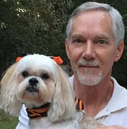 An older man with a light beard smiling gently while holding a small white dog with a fluffy head, both featuring matching orange bows. They seem to be outdoors with trees in the background, near a veterinarian's clinic.