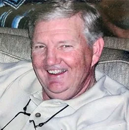 A smiling older man with light hair wearing a beige shirt, seated on a sofa with blue patterned cushions in the background, is a retired veterinarian.